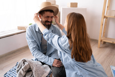 a young cheerful couple in a new apartment on the day of the move sit on the floor unpack things the woman puts on a hat to the man. moving to a new apartment. Lots of packing boxes around them.の写真素材