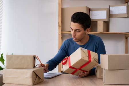 a working Muslim man in an online store. He wears casual clothes and packs goods for delivery. Muslim small business owners, packing goods into boxes, preparing them for deliveryの写真素材