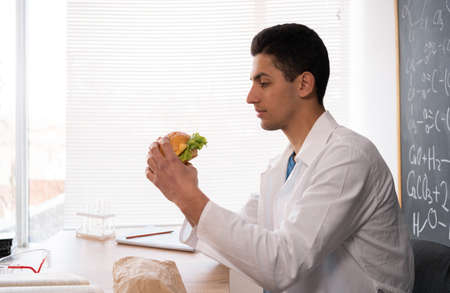 Young teacher having breakfast with hamburger at workplace, lunch in classroom with junk cheeseburger, student in white coat indoors eating fast food, nutrition conceptの写真素材