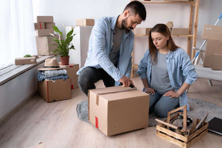 A man and a woman on the day of the move sit on the floor and unpack things. A man cuts the adhesive tape on a cardboard box with a knife. Happy couple in a new apartment. copyspace.の写真素材