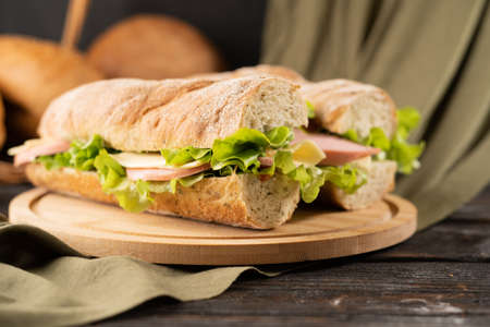 fast food concept, Two fresh submarine sandwiches with ham, cheese, bacon, tomatoes, lettuce, cucumbers and onions on wooden cutting boardの写真素材