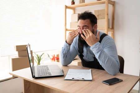 tired indian male small business owner working at laptop, online store delivery worker has vision problems, businessman working at home office, sme conceptの写真素材