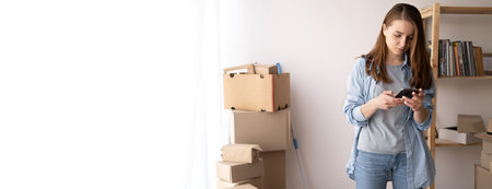 serious young woman is moving, standing among cardboard boxes, using a smartphone, moving day and...