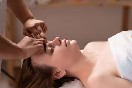Beautiful woman enjoying facial massage with closed eyes, close up of masseuse hands massaging female face.の写真素材