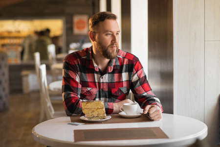 Young bearded man drinking coffee with a cake in the morning in a cafe, a businessman starting his working day with breakfast in a restaurantの写真素材