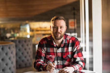 smiling bearded cafe owner planning work. male restaurant chef sits at the table after hours doing her bookkeeping in front of an camera. student search new idea inspiration in office cafeの写真素材
