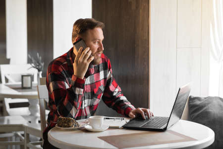 Successful businessman talking on smartphone in cafe, working in cafe during coffee break, man using modern laptop in stylish cafe. Casual man making notes while working on laptop computerの写真素材