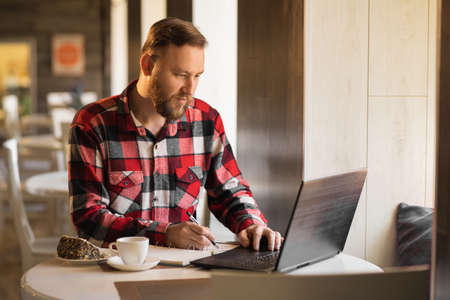 businessman working with documents and laptop in cafe. bearded blogger in plaid shirt taking notes in cafeteriaの写真素材