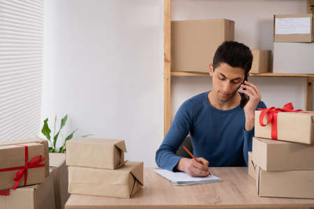 muslim male salesman confirming orders from a customer by phone makes notes on paper. An e-commerce arabic business owner looks at documents and speaks on the phone in a warehouse.の写真素材