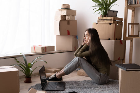 Attractive young woman is moving, sitting among cardboard boxes, using a laptop and smiling...
