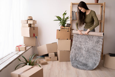 Young latin woman moving to a new house, collecting carpet and cardboard boxes with things for relocation, moving dayの写真素材