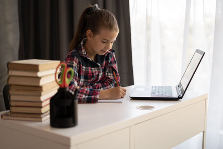 Little student sit at desk at home look at laptop screen studying and making notes, concentrated...