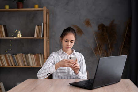 Young Hispanic student holding smartphone studying on laptop, using mobile learning classes tech apps. Teenage girl watching online training at office.の写真素材