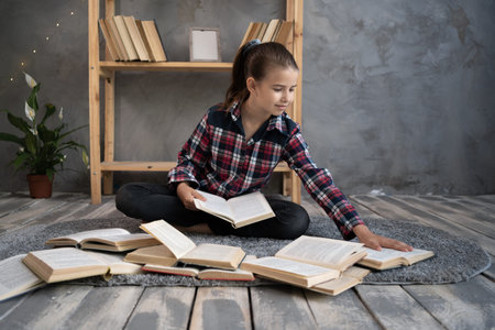 schoolgirl is sitting on the floor library with stacks of books beside. Close-up, pupil holding read...