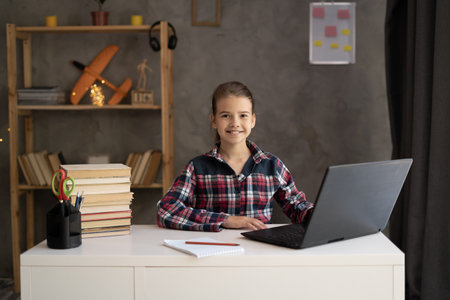 Cute smart Caucasian schoolgirl virtual distance learning online, pupil watching remote digital class lesson looking at laptop computer tech studying at home writing notesの写真素材