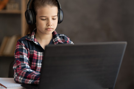 Portrait of little girl in wireless headset using laptop, studying online at home, student typing on keyboard looking at pc screen, watching webinarの写真素材