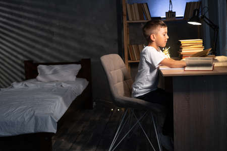 Young boy studying at desk in bedroom in evening, schoolboy at the table doing homework in the dark room, child learning at nightの写真素材