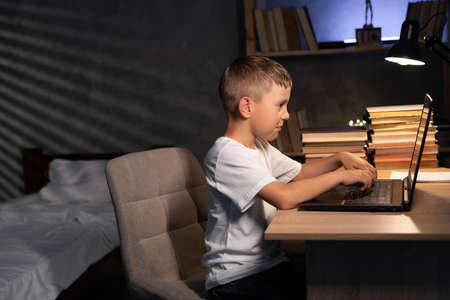 Caucasian boy studying at night at home using laptop computer sitting at table, schoolboy learning online, e-learningの写真素材