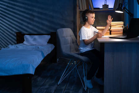 focused schoolboy wearing headphones uses laptop for distance learning at night. child raises his hand to answer lesson. e-education concept. Distance learningの写真素材