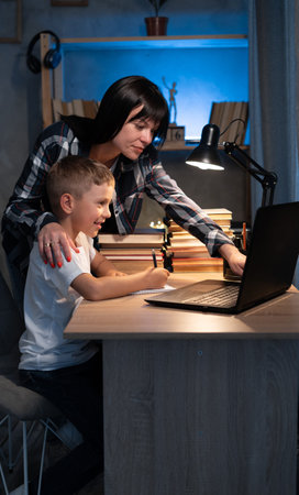 Cute boy with mother doing homework at home using laptop and books at night, online schooling
