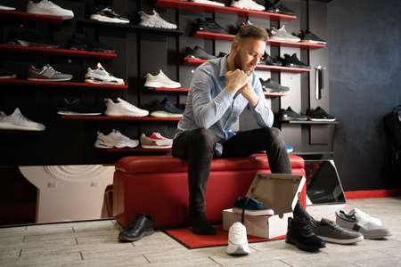Cheerful man chooses shoes in a sports shop while sitting in fitting room of modern storeの写真素材