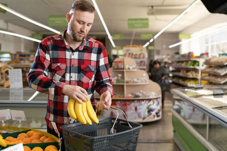 Grocery shopping. Cheerful man in supermarket choosing fresh fruits and vegetables, carrying basket buying natural healthy food in store.の写真素材