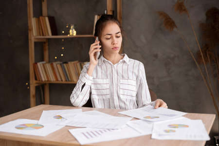 serious Hispanic young business woman with papers working at home office and calling on smartphone. remote job, technology and peopleの写真素材