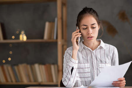 young hispanic woman with papers working at home office and calling on smartphone, serious businesswoman checking letter calling on sell phone sitting in the living room at homeの写真素材