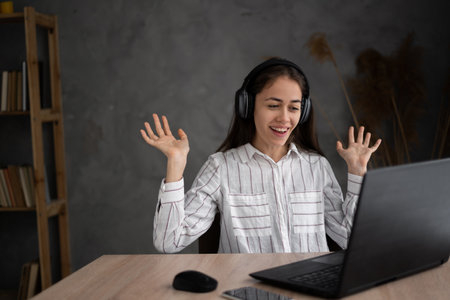 happy young hispanic business woman in casual shirt headset for helpline assistance working at office desk with pc laptop spreading hands on background portraitの写真素材