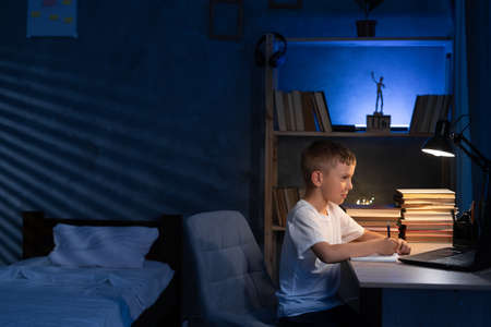 Boy studying at home at night using laptop, student doing homework late evening with computer, online lesson, e-learning, modern learning technologies and teachingの写真素材