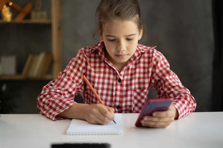 Concentrated child girl doing homework at the her room, student learning at home using smartphone for remote education with online school teacher or personal tutor. Homeschoolingの写真素材