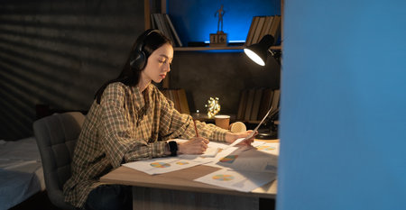 Overworked. Asian female student working on project, checking financial documents, sitting at table at home at late night, looking at paperwork with diagramsの写真素材