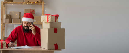 Indian business owner Santa Claus working with product packaging and smartphone wearing a hat and red sweater. Calling phone and prepares gifts purchase for shipment. Christmas saleの写真素材