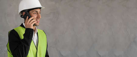 Young architect dressed in protective helmet is working in construction area. Foreman use smartphone on cement wall with copy space, bannerの写真素材