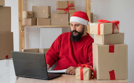 Indian small and medium business freelance Santa working in home office using computer, online marketing packaging box and gifts for Christmas delivery, SME e-commerce telemarketingの写真素材