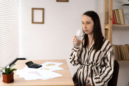 Pregnant woman drinking water and working at home office, pregnant freelancer holding glass of water in officeの写真素材