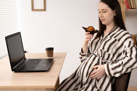 pregnant woman is working on computer laptop and eating delicious cupcake, lunch for mother, business, food and pregnancyの写真素材