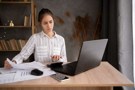 Freelance young hispanic businesswoman using laptop working at home with paper documents and charts. Working from home, remotely work, self isolationの写真素材