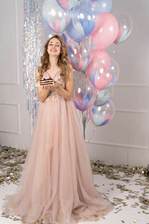 Happy Birthday. Young woman with balloons and cake at party. Celebrating holiday. Portrait of beautiful smiling girl in festive dress holding cakeの写真素材