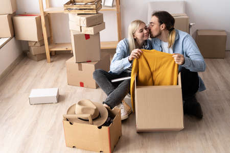 Young couple moving into a house, unpacking boxes while sitting in a new apartment on the floor, clothes from volunteersの写真素材
