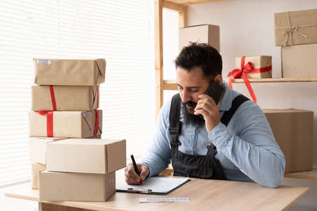 Indian bearded man online seller confirming orders from customer on the phone. E-commerce male business owner using phone in store warehouse. SMEの写真素材