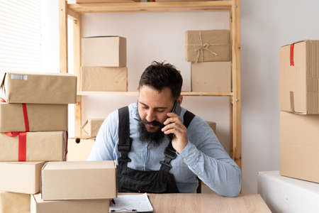 bearded man online seller confirming orders from customer on the phone. E-commerce male business owner using phone in store warehouse with boxes for shipping. SMEの写真素材