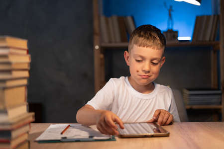 Boy using digital tablet while sitting on desk at home at night. Reading and studyingの写真素材