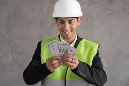 Young arabic builder man engineer wearing safety helmet holding dollars as payment over background with raised hand, payment constructionの写真素材