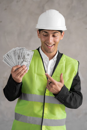 Young muslim builder man engineer wearing safety helmet holding money dollars as payment over background, a foreman showing cash with an expression of shock, payment work constructionの写真素材