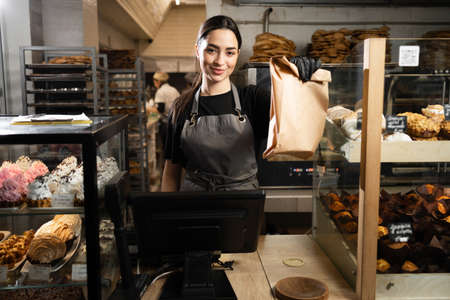baker woman standing with fresh bread and pastry at bakery. Young woman in her bake shop looking at camera. baker with breads in background. Girl owner bakery shopの写真素材