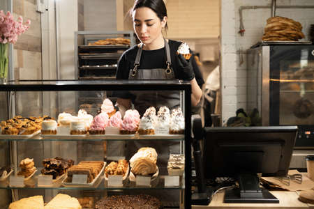 Handsome woman coffee shop employee placing pastry and cake in bakery refrigerator showcase at cafe. Small business, part time jobの写真素材