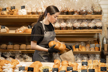 Young smiling girl with bread in the bakery, preparing pastry for sale in supermarket bakery department.の写真素材