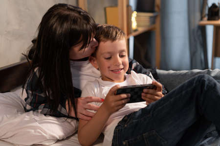 Mother and son woman and her boy child sitting on the bed at home making video call or playing video games on mobile smart phone at home in eveningの写真素材