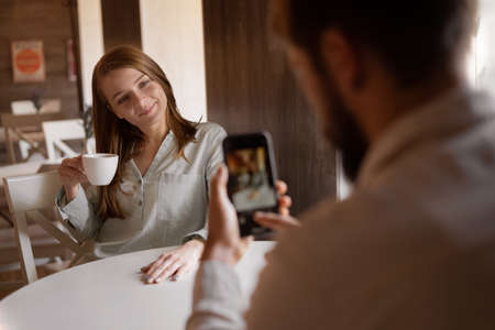 couple in love sitting in cafe, male photographer photographing woman on smartphone with a cup of coffee , indoor mobile photography, Social media blog introduction conceptの写真素材